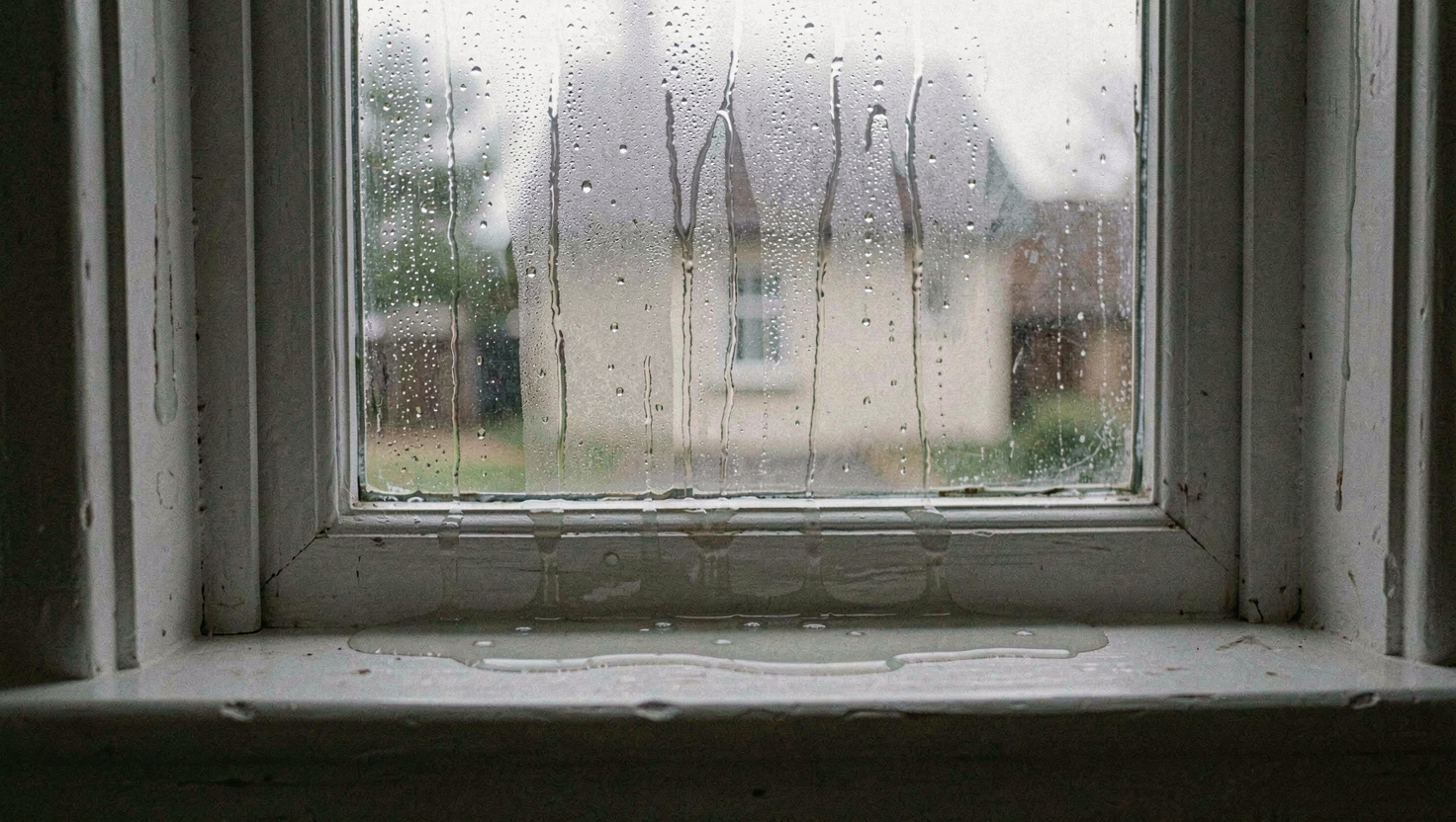 Heavy condensation running down the inside of a single glazed sash window with a pooled line of water on the painted sill, grey morning light behind, close shot across the glass