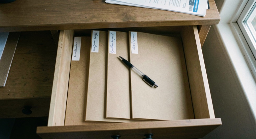 An open desk drawer holding four upright manila folders with handwritten tabs, a pen resting across them, cool daylight from a window on the right, shot from directly above