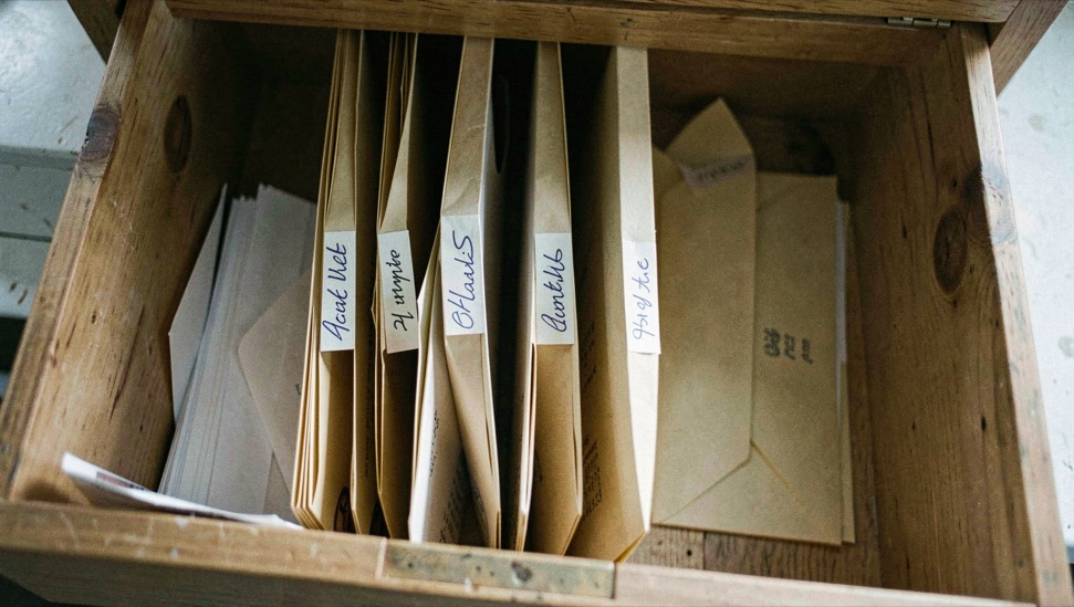 A wooden desk with an open folder of copied certificate pages, a photograph of a stone war memorial and a pencil beside it, window light from the right, photographed from above
