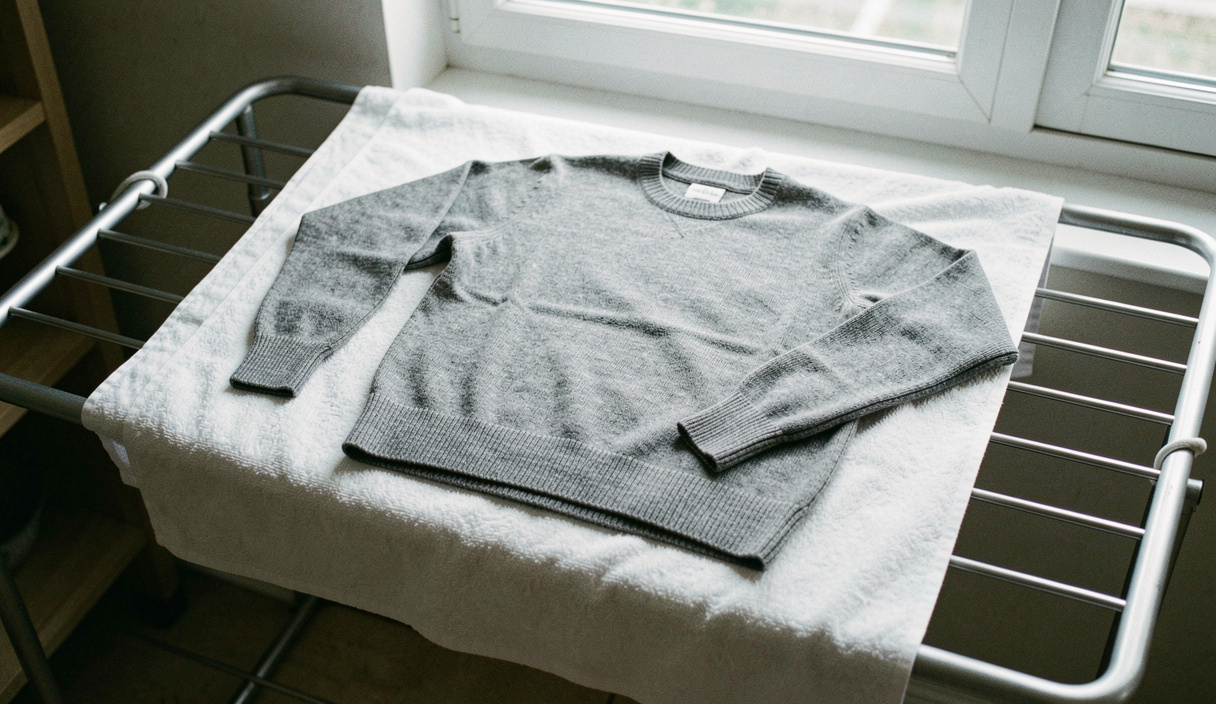 A grey wool jumper laid flat to dry on a white towel across a slatted rack, sleeves squared, cool daylight from a window behind, photographed from above at an angle